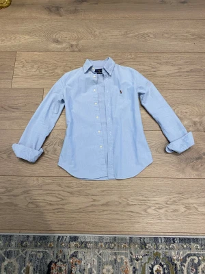 Ljusblå skjorta från Ralph Lauren - Klassisk ljusblå skjorta från Ralph Lauren med broderad logga på bröstet. Skjortan har lång ärm, knappar framtill och en tidlös krage. Perfekt för en clean och stilren look.