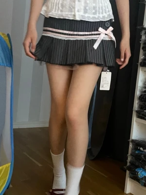 Grå lågmidjad miniskirt med rosett och spets - Säljer en grå lågmidjad minikjol med vita smala ränder, rosa band och en söt rosett framtill. Kjolen har plisserad nederkant och spetsdetalj. Perfekt för dig som gillar en gullig och trendig stil.