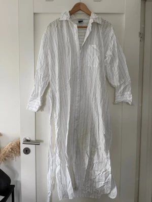 Vit lång skjortklänning med svarta ränder - Vit lång skjortklänning med tunna svarta vertikala ränder, klassisk krage och lång ärm. Knäppning framtill och bröstficka. Tillverkad i mjuk bomull, perfekt för en avslappnad och stilren look. Fästet för bältet har lossnat på ena sidan, köpte den så- inget jag gjort!!