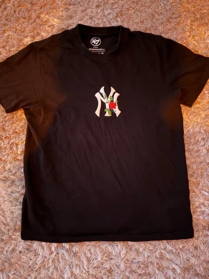 Svart t-shirt från 47 Brand - Svart t-shirt från 47 Brand med New York Yankees-logga och broderad ros på bröstet. Klassisk passform och rund halsringning. Tillverkad i 100% bomull för en skön känsla. Perfekt för dig som gillar sportig streetstyle.