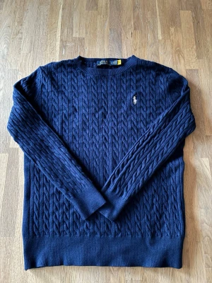  Ralph Lauren sweater navy blå - Snygg mörkblå kabelstickad tröja från Polo Ralph Lauren med klassisk rund halsringning och broderad logga på bröstet. Tröjan har långa ärmar och ribbade muddar. Perfekt för dig som gillar stilrena och tidlösa plagg.