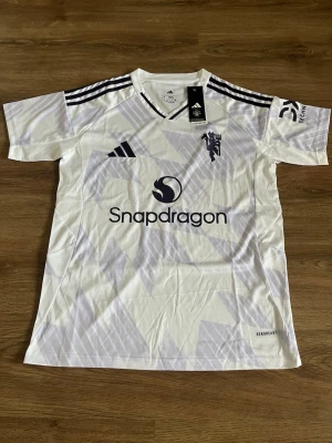 Vit Manchester United matchtröja Adidas - Snygg vit Manchester United matchtröja från Adidas med svarta detaljer och tre ränder på axlarna. Tröjan har tryckt Snapdragon-logga på bröstet och klubbmärke på vänster sida. Tillverkad i lätt och ventilerande material, perfekt för träning eller match.