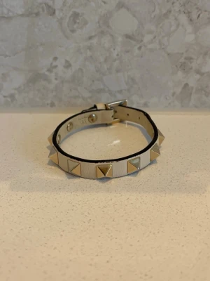  valentino armband - Snyggt beige armband i läder med silverfärgade pyramidformade nitar runt om. Justerbart spänne för perfekt passform. Perfekt accessoar för dig som gillar en edgy och trendig stil.