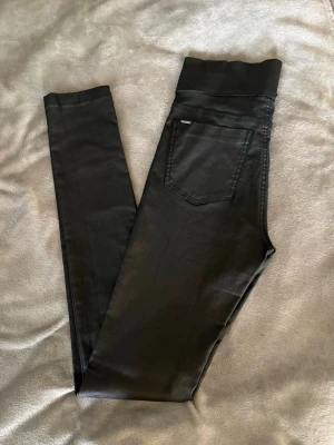Svarta coated leggings med dragkedjor - Snygga svarta leggings med coated finish som ger en läderlook. Bred resår och två dragkedjefickor fram. Bakficka som detalj. Stretchigt material för skön passform och trendig stil. Står ingen storlek men skulle gissa S