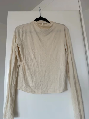Beige långärmad polotopp från Nakd - En enkel och stilren beige polotopp med lång ärm. Toppen har en rak passform och är tillverkad i mjukt material som känns skönt mot huden. Perfekt att matcha med jeans eller kjol för en clean look.