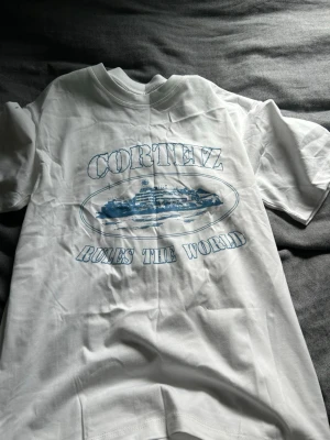 Vit Corteiz t-shirt med blått tryck - Vit t-shirt från Corteiz med blått tryck på bröstet där det står 'Corteiz Rules The World' och en grafisk bild av ett fartyg. Klassisk rund hals och korta ärmar. Tillverkad i mjuk bomull för skön känsla.