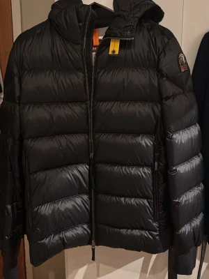 Svart dunjacka från Parajumpers - Snygg svart dunjacka från Parajumpers med huva och dragkedja framtill. Jackan har breda, quiltade paneler och en gul detalj vid halsen. Märkespatch på ärmen och praktiska fickor. Perfekt för kalla vinterdagar.