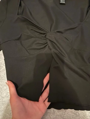 Svart croppad tröja - Snygg svart croppad topp med lång ärm. Toppen har en twistad detalj framtill och en djup v-ringning som ger en trendig look. Tillverkad i ett stretchigt syntetmaterial som sitter tight och bekvämt.