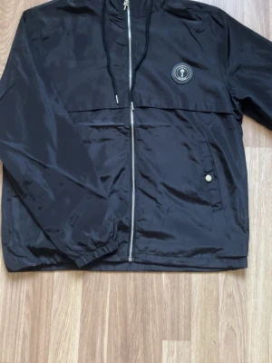 Trapstar Svart windbreaker - Snygg svart vindjacka med hel dragkedja framtill, huva med snören och en ficka med knapp på sidan. Jackan har ett runt märke på bröstet och är tillverkad i ett glansigt, tunt material som passar perfekt för blåsiga dagar.