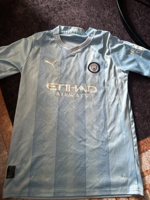 Manchester City matchtröja Puma - Ljusblå Manchester City matchtröja från Puma med klubbmärke, Etihad Airways-tryck och nummer 7 samt namnet Ramzi på ryggen. Tillverkad i lätt och ventilerande material, perfekt för fotboll eller träning. Klassisk V-ringning och korta ärmar.