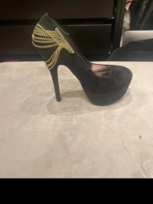 Svarta pumps med guldkedjor - Snygga svarta pumps med hög klack och platåsula. Dekorerade med flera guldfärgade kedjor på hälen som ger en unik och trendig look. Skorna har en spetsig tå och är tillverkade i mockaliknande material.