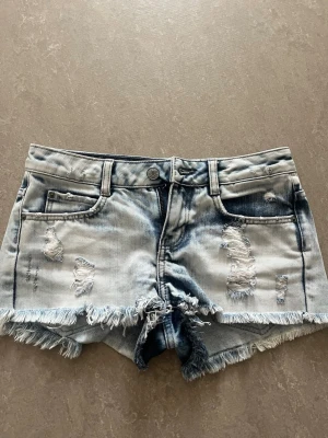 Ljusblå slitna jeansshorts med fransar - Säljer ett par ljusblå jeansshorts med slitna detaljer och fransig kant. Shortsen har fem fickor, bälteshällor och knappstängning. Perfekta för sommaren och ger en avslappnad streetstyle-vibe.