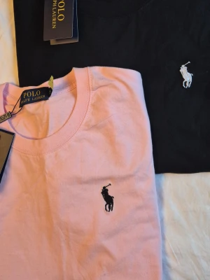 2-pack Polo Ralph Lauren t-shirts - Säljer två klassiska t-shirts från Polo Ralph Lauren, en i ljusrosa och en i svart. Båda har rund hals, korta ärmar och den ikoniska broderade loggan på bröstet. Tillverkade i mjuk bomull för en skön och stilren look.