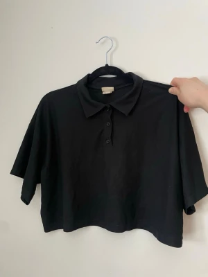 Svart croppad polotopp med knappar - Snygg croptop som kommer från H&Ms tenniskollektion. Går både att träna och dansa i. Bra längd, bekväm och ligger skönt på kroppen. Lägger sig snyggt på axlarna.  Sparsamt använd.