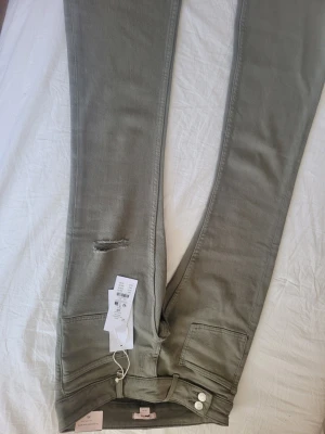 Grågröna skinny jeans från Gina Tricot - Snygga grågröna skinny jeans från Gina Tricot med slitning vid låret. Jeansen har hög midja, klassiska fem fickor och stängs med två silverfärgade knappar. Materialet är stretchigt för en bekväm passform. Perfekta till en avslappnad look.