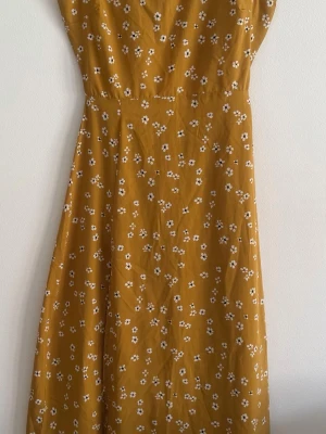 Gul blommig långklänning med cut out - Säljer en gul långklänning med små vita blommor och öppen rygg. Klänningen har en figurnära midja och ett snyggt cut out-detalj bak. Perfekt för varma dagar och sommarens alla tillfällen.