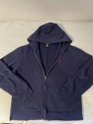 Mörkblå hoodie från Ralph Lauren - Mörkblå hoodie med dragkedja från Ralph Lauren. Klassisk design med huva, två fickor framtill och den ikoniska röda loggan broderad på bröstet. Tillverkad i mjukt bomullsmaterial som känns skönt mot huden. Perfekt för en avslappnad och stilren look. Tröjan är XL i barn storlek men S/M i men