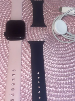 Apple Watch - Säljer en Apple Watch med en rosa silikonrem och ett extra svart silikonarmband. Klockan har en fyrkantig svart skärm och kommer med en magnetisk laddare med USB-C. Perfekt för dig som vill ha koll på både träning och notiser direkt på handleden. Kostar 2700 ny. Mitt pris 1000kr