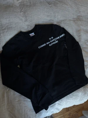 Svart Supreme x Comme des Garçons tröja - Supreme x Comme des Garçons svart långärmad tröja med vit text på bröstet. Klassisk crewneck-modell i mjuk bomull, perfekt för dig som gillar streetwear och vill sticka ut med en limiterad collab. Helt ny❤️