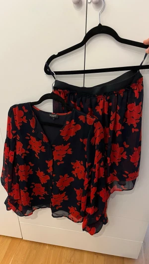 Blommigt set från Soaked in Luxury - Snyggt tvådelat set från Soaked in Luxury med transparent svart tyg och stora röda blommor. Setet består av en långärmad blus med ballongärmar och en matchande kjol med resår i midjan. Luftigt material som ger en cool och trendig look. Storlek S i blusen och M i kjolen.