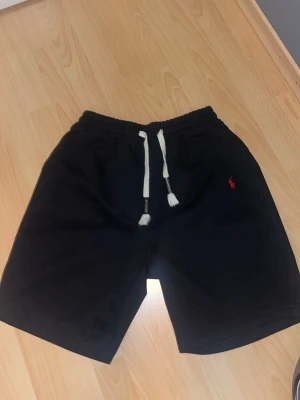 Svarta shortsRalphLauren - Svarta shorts från Polo Ralph Lauren med vit snörning i midjan och den klassiska röda loggan broderad på benet. Mjukt material och avslappnad passform, perfekta för sommardagar eller chill hemma. Helt nya storlek XL