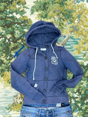 Blå hoodie med knappar från Hollister - Mysig blå hoodie från Hollister med knappar framtill och vit broderad logga på bröstet. Tröjan har huva med vita dragsnören och ribbade muddar vid ärmslut och nederkant. Perfekt för chill dagar och enkel att matcha med jeans.