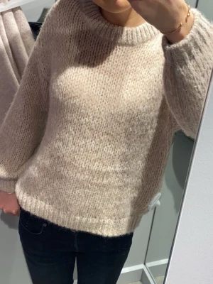 Beige stickad tröja - Mysig beige stickad tröja med rund hals och långa ärmar. Tröjan har en fluffig, lite luddig struktur och ribbade muddar vid ärmslut och nederkant. Perfekt för lager på lager och en avslappnad look. 
