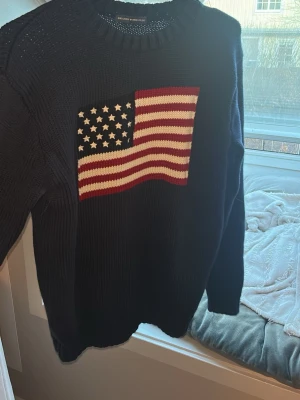 Mörkblå stickad tröja Brandy Melville - Stickad tröja i mörkblått från Brandy Melville med amerikansk flagga i rött, vitt och blått på bröstet. Tröjan har rund halsringning och ribbade ärmar och nederdel. Jätte mysig och bra passform! 