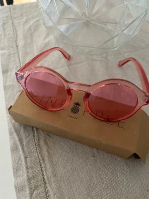 Rosa runda solglasögon Pineapple - Säljer ett par riktigt snygga solglasögon från Pineapple med rund form och transparent rosa båge samt rosa glas. Perfekta för att ge din look en färgklick och cool retrovibe. Kommer med originalkartong.