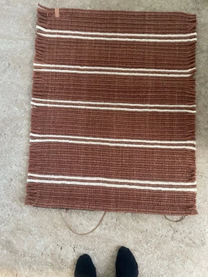 Brun handvävd matta med ränder - Säljer en fyrkantig handvävd matta i brun färg med vita horisontella ränder. Mattan har en rustik känsla och fransar på kortsidorna. Passar perfekt som inredningsdetalj i sovrum eller hall.