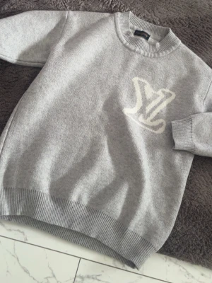 Grå stickad tröja från Louis Vuitton - Snygg grå stickad tröja från Louis Vuitton med vit LV-logga på bröstet. Tröjan har rund halsringning och ribbade muddar vid ärmslut och nederkant. Perfekt för dig som gillar exklusiva och stilrena plagg.