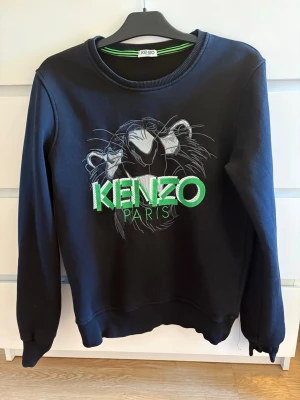 Svart sweatshirt från Kenzo - Cool svart sweatshirt från Kenzo med ikoniskt tigerbroderi och grönt Kenzo Paris-tryck på bröstet. Tröjan har rund halsringning, långa ärmar och ribbade muddar. Perfekt för dig som vill ha en streetwear-vibe och sticka ut med ett snyggt märke.