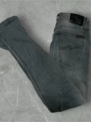 Grå jeans från Nudie Jeans - Snygga grå jeans från Nudie Jeans med klassisk femficksmodell och svarta detaljer på bakfickan. Jeansen har en rak passform och är tillverkade i slitstark denim. Perfekta för en avslappnad och trendig look.