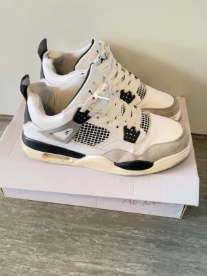 Vita och svarta Air Jordan 4 sneakers - Säljer ett par Air Jordan 4 sneakers i vitt och svart med grå detaljer. Skorna har meshpaneler på sidorna, klassiska plastdetaljer och synlig Air-sula. Perfekta för dig som gillar streetwear och vill ha en ikonisk look.