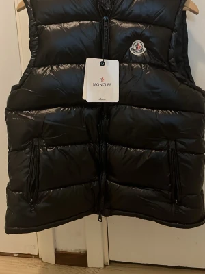 Svart dunväst från Moncler - Snygg svart dunväst från Moncler med glansig finish, hög krage och dragkedja framtill. Västen har två stora fickor med dragkedja och Moncler-logga på bröstet. Insidan har ett unikt serietidningsmönster och etiketter med info om material och skötsel.