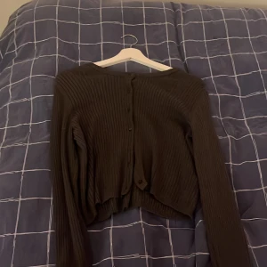 Svart ribbad kofta med knappar - Säljer en svart ribbad kofta med långa ärmar och knäppning bak. Koftan har en croppad passform och är tillverkad i ett mjukt, stretchigt material. Perfekt att styla över ett linne eller t-shirt för en chill look.