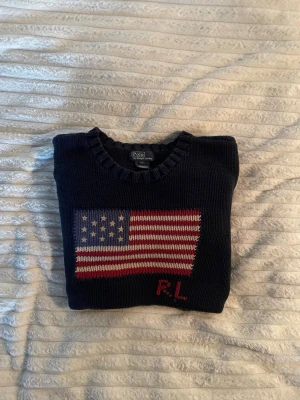 Mörkblå stickad tröja Polo Ralph Lauren - Mörkblå stickad tröja från Polo Ralph Lauren med amerikansk flagga i rött, vitt och blått på bröstet samt RL-broderi i rött. Tröjan har rund halsringning och ribbstickade muddar vid ärmslut och nederkant. Perfekt för dig som gillar klassisk preppy stil.
