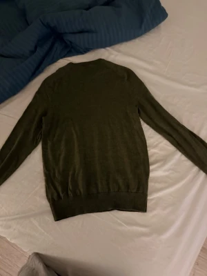 Olivgrön stickad tröja från Morris - Snygg olivgrön stickad tröja från Morris med rund halsringning och diskret logga broderad på bröstet. Tröjan har långa ärmar och ribbade muddar vid ärmslut och nederkant. Perfekt för lager på lager och enkel att matcha. Det är 100% merino ull