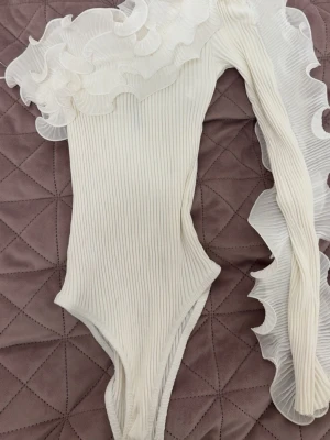 Vit body med volangdetaljer - En vit ribbad body med långa ärmar och stora volangdetaljer längs axel och ärm. Dessutom finns det knappar för att få av och på sig