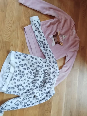 Två mjuka tröjor med glitterdetaljer - Säljer två långärmade tröjor. En ljusrosa tjock-tröja med paljettstjärna i guld och silver på bröstet, samt en vit tjock-tröja med grått leopardmönster. Båda är i mjukt material som en filt. Älskade de här när jag var 9 år! (Men det märks inte att de är välanvända)