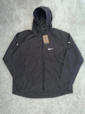Helt ny svart vindjacka från Nike med huva - Snygg svart vindjacka från Nike med dragkedja framtill och justerbar huva. Jackan har en vit Nike-logga på bröstet och är tillverkad i lätt, vindtätt material. Perfekt för blåsiga dagar och sportiga outfits. Storlek XL men är ganska liten i storlek så jag skulle säga att den passar M