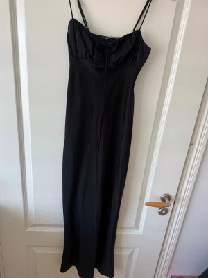 Svart långklänning från Gina Tricot - Snygg svart långklänning från Gina Tricot med smala axelband och snörning vid bysten. Klänningen har en figurnära passform och är tillverkad i ett mjukt, lätt material som ger en elegant siluett. Perfekt för dig som vill ha en stilren och trendig look.