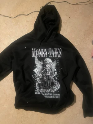 Svart hoodie med Money Talks-print - Svart hoodie med stor grafisk print på framsidan som föreställer en ängel med pengar och texten 'Money Talks'. Hoodien har huva och en avslappnad passform. Perfekt för dig som gillar streetwear och statement-plagg.