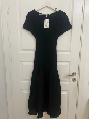 Svart långklänning från H&M - Elegant svart långklänning från H&M med korta ärmar och smockad överdel. Klänningen har en mjuk, flytande kjol och rundad halsringning. Perfekt för dig som gillar en klassisk och stilren look.