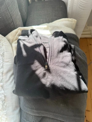 Svart fleecehoodie med halv dragkedja - Mjuk och varm svart fleecehoodie från Helly Hansen med halv dragkedja framtill och huva. Hoodien har en diskret logga på ärmen och är tillverkad i skönt fleecematerial som passar perfekt för kyliga dagar.