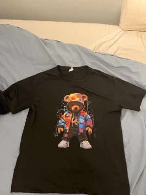 Svart t-shirt med färgglad björn - Svart t-shirt med en stor tryckt björn på framsidan. Björnen har en färgglad hoodie och sneakers, vilket ger en cool streetwear-vibe. T-shirten har rund hals och korta ärmar, perfekt för en avslappnad stil.