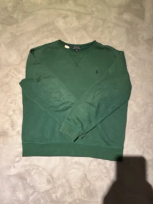 Grön sweatshirt från Polo Ralph Lauren - Mörkgrön sweatshirt från Polo Ralph Lauren med rund halsringning och klassisk broderad logga på bröstet. Tröjan har ribbade muddar vid ärmslut och nederkant samt en avslappnad passform. Perfekt för en chill och stilren look.