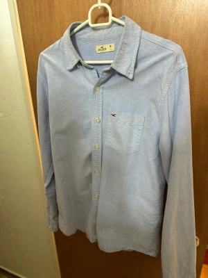 Ljusblå skjorta från Hollister - Säljer en ljusblå skjorta från Hollister med klassisk krage och bröstficka med liten logga. Skjortan har långa ärmar och knäppning framtill. Perfekt för en clean och avslappnad stil.