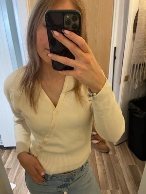 Krämvit ribbad kofta med v-ringning - Snygg krämvit kofta med v-ringning och ribbade muddar. Koftan har långa ärmar och stängs med flera matchande knappar framtill. Perfekt att styla med jeans för en clean och enkel look.