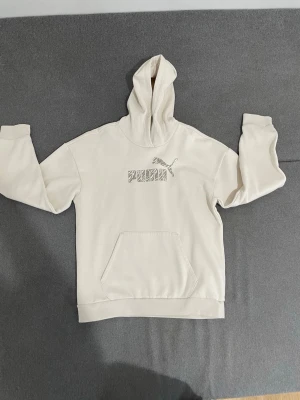 Beige hoodie från Puma med logga - Snygg beige hoodie från Puma med stor logga och zebramönstrad text på bröstet. Klassisk känguruficka framtill och ribbade muddar vid ärmar och nederkant. Tillverkad i mjukt material och har en stor huva för extra komfort.